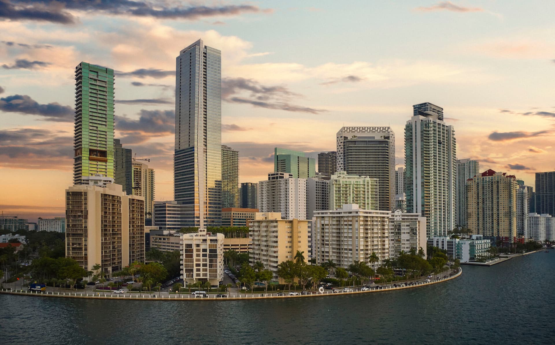 Drone perspective over Biscayne Bay of curved shoreline, hotels, condominiums, and skyscrapers beneath moody sky.