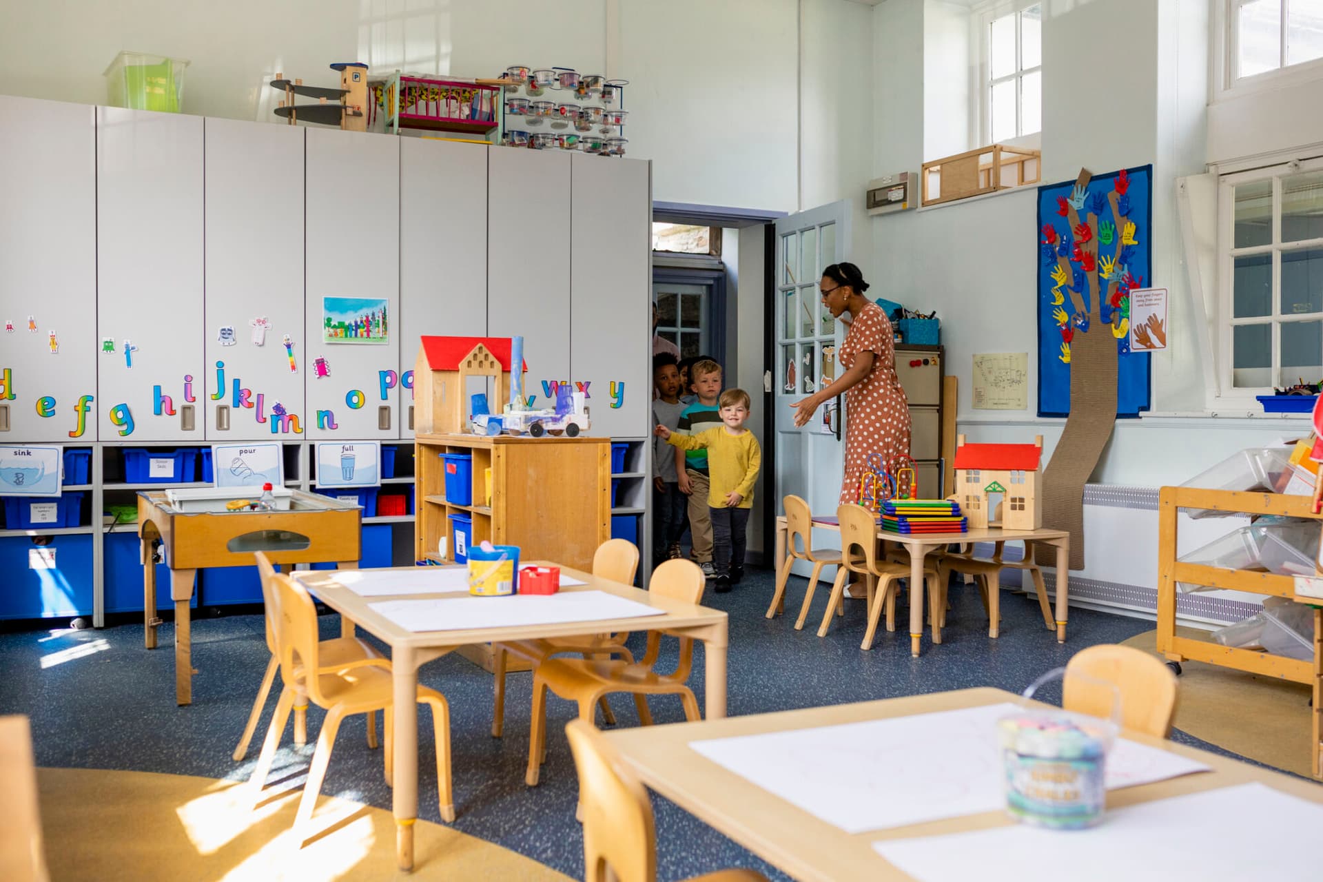 Primary school students coming into an art classroom at school. The teacher is welcoming them in.