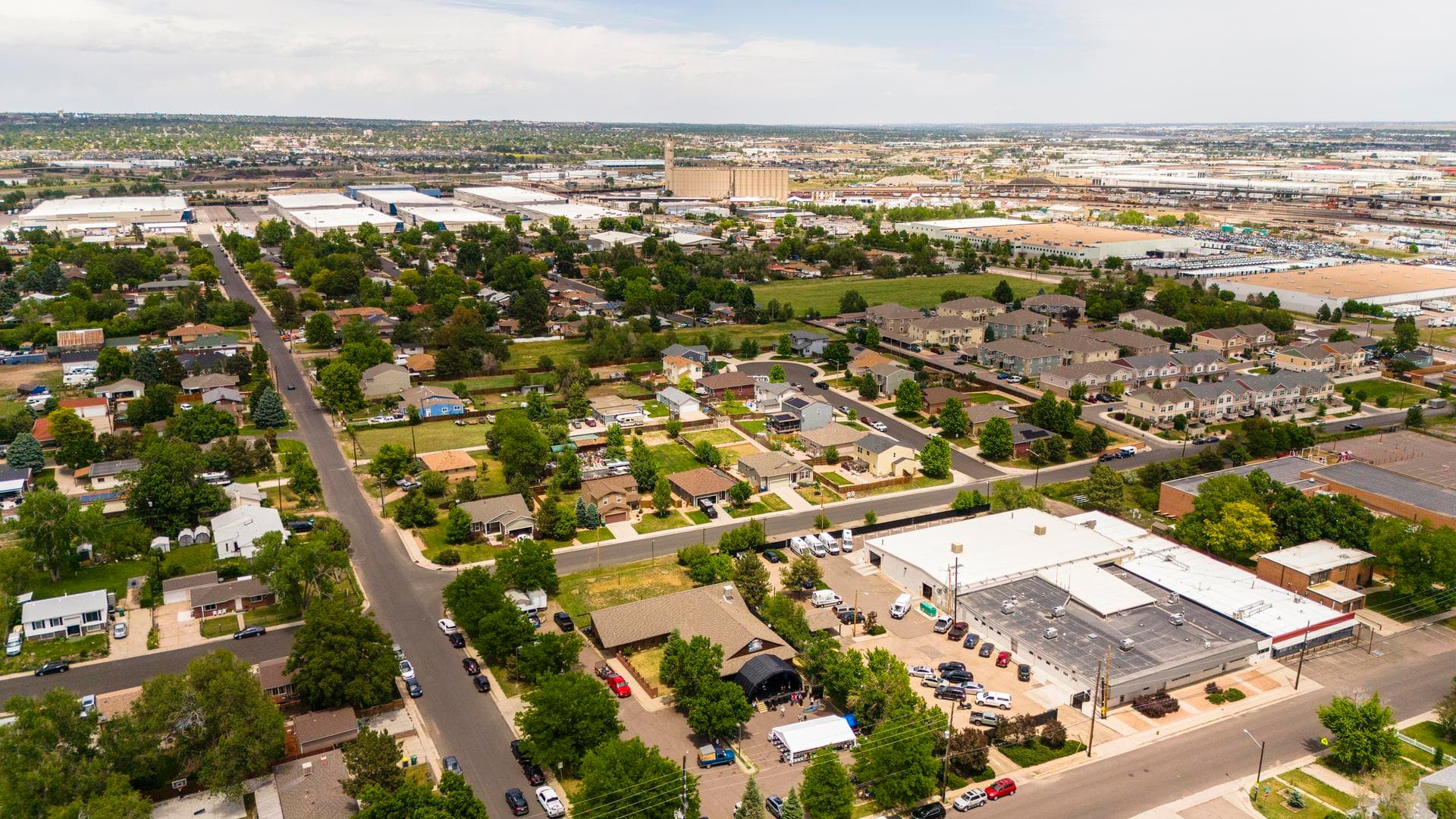 Aerial view showing transition between residential neighborhood and industrial district during summer afternoon in Denver, Colorado. Single family houses with green lawns sit adjacent to large commercial buildings and warehouses. A grain elevator is visible in the distance under a partly cloudy sky.